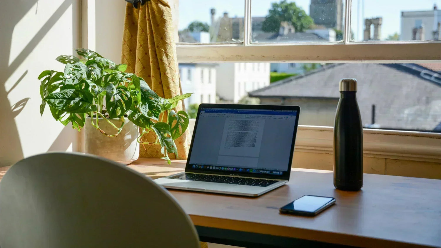 Postazione di lavoro smart working con laptop aperto su una scrivania in legno vicino a una finestra luminosa, con una pianta verde Monstera, una borraccia nera e uno smartphone.